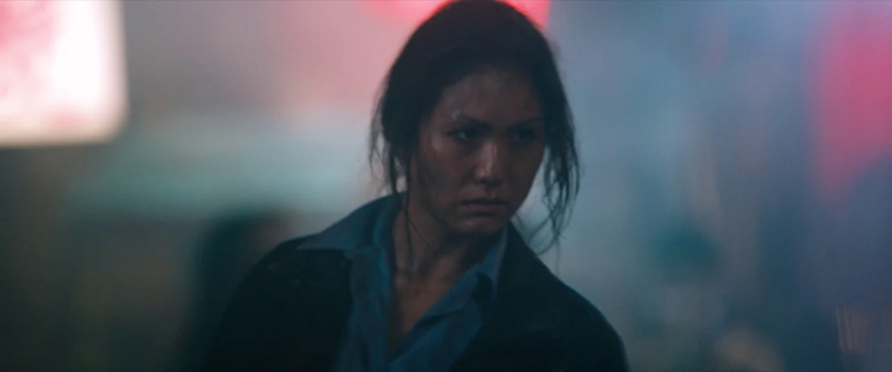 Venom (2018) movie still: closeup — A dimly lit city street at night filled with heavy atmospheric smoke and haze. An East Asian woman with damp…