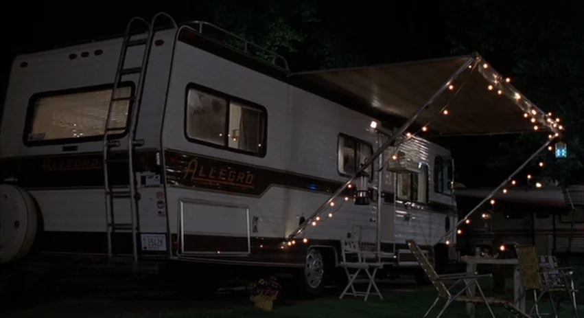 About Schmidt movie still: wide — A white Allegro motorhome is parked at a campsite during the dark night hours. String lights are draped under an…
