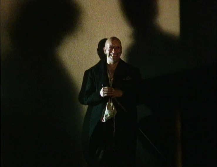 The Tempest (1979) movie still: medium — A dark indoor space with a textured wall lit by a high-contrast source. A bald man wearing an overcoat…