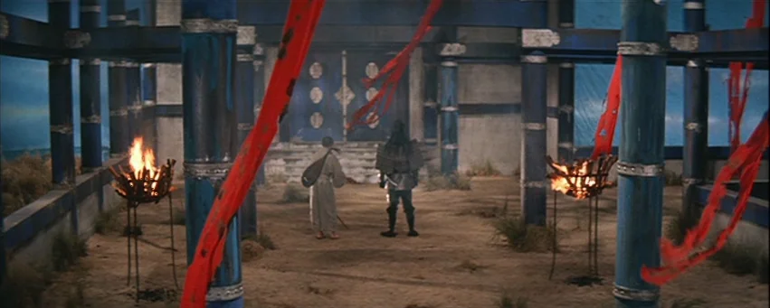 Kwaidan (1964) movie still: wide — A spacious hall with sandy ground, blue pillars, and draped red ribbons hanging overhead. Two men stand facing a…