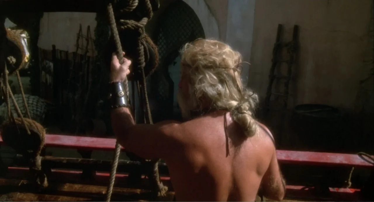 Hercules (1983) movie still: medium — The deck of a wooden sailing ship features ropes, pulleys, and a wooden railing. A muscular man with blonde…