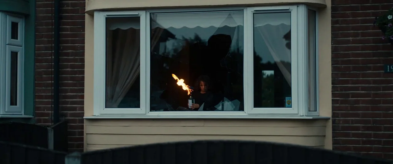 Silver Haze (2023) movie still: wide — A suburban brick house features a bay window with multiple shattered glass panes. A young boy stands inside…