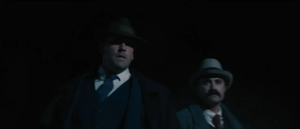 Fear Below (2025) movie still: medium — Two men wearing period-appropriate hats and coats stand in a dark, outdoor space. They walk forward together…