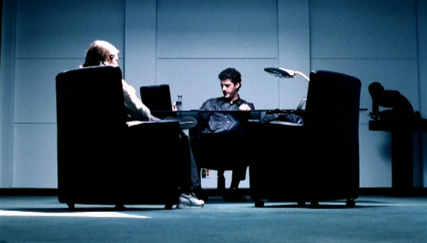 Maelström (2000) movie still: wide — A sterile room features a long glass desk positioned against a flat wall. Two men sit in high-backed chairs…