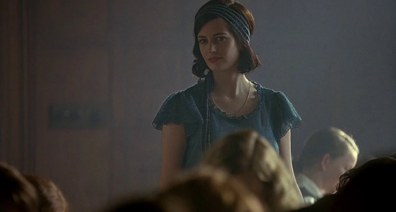 Cracks (2009) movie still: medium — A dimly lit room with dark wood paneling and blurred heads in the foreground. A woman with dark hair stands in…