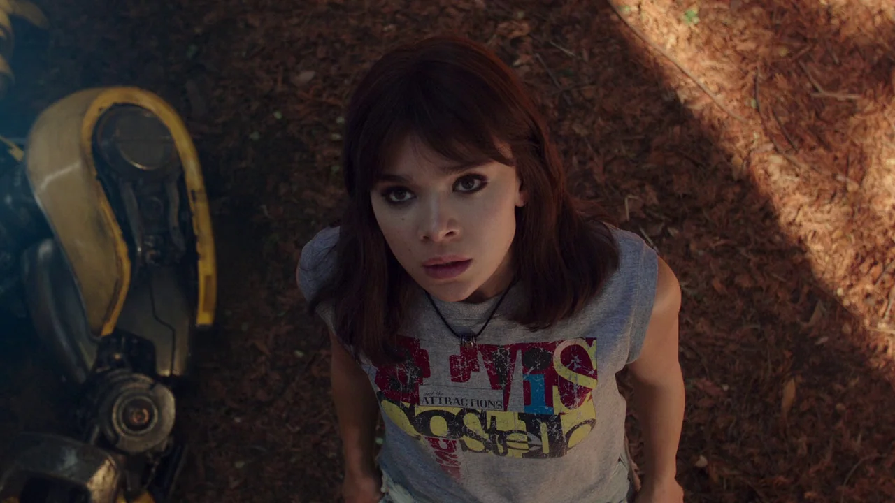 Bumblebee (2018) movie still: medium — A clearing with a bed of dark dry leaves on the ground. A young woman stands looking upward toward the camera…