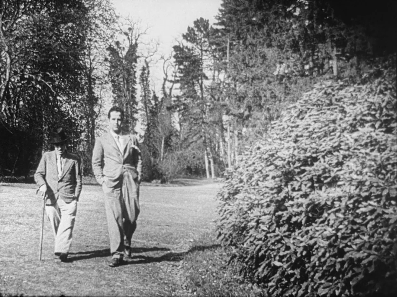 Un Chien Andalou (1929) movie still: fullbody — A wide park path is lined by tall mature trees and a large bush. Two men walk side-by-side along the…