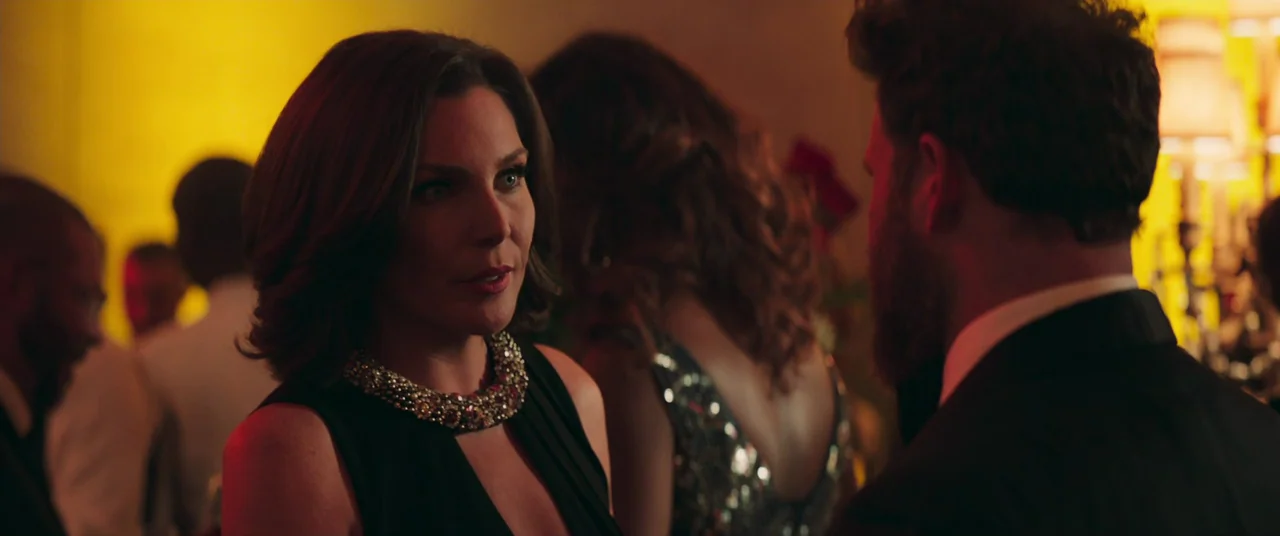 Long Shot (2019) movie still: medium — A crowded social gathering interior with guests dressed in formal attire under dim lighting. A woman engages…