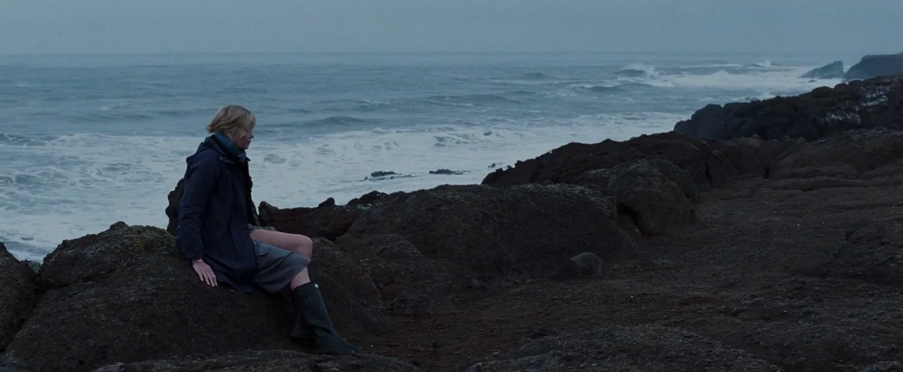 The Burning Plain (2008) movie still: wide — A dark rocky shoreline sits beside a turbulent gray sea under an overcast sky. A woman in a blue…