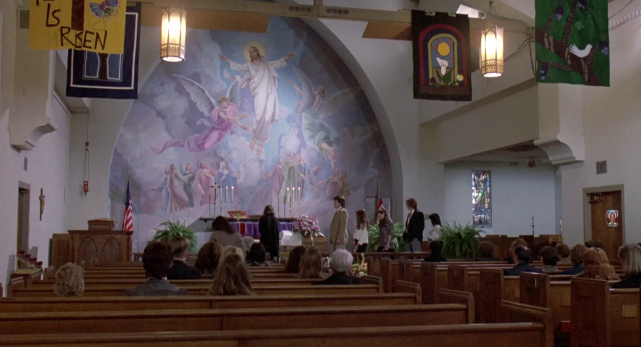 River’s Edge (1986) movie still: wide — A church nave features rows of wooden pews facing a large mural of Jesus. People stand and walk near the…