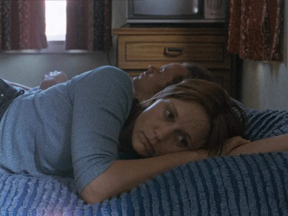 River of Grass (1994) movie still: medium — A woman and a man lie together on a bed with a textured blue spread. An old television set sits on a…