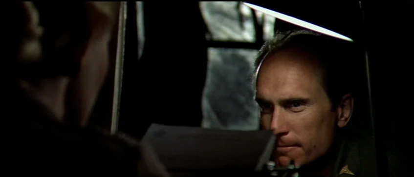 MASH (1970) movie still: closeup — A dark, dimly lit office space with a subject illuminated by a desk lamp. The subject faces across a desk toward…