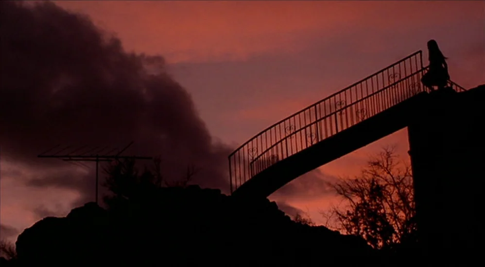 U-Turn (1997) movie still: wide — A metal footbridge stands against a deep red and purple sunset sky. A lone figure stands on the bridge while a…
