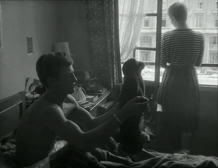 À Bout De Souffle (Breathless) (1960) movie still: medium — A spartan bedroom with a wooden bed frame and a large windowed wall. A man sits…