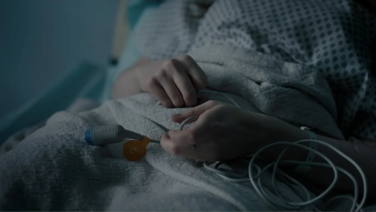 Sightless (2017) movie still: closeup — A hospital bed with a white patterned gown and medical equipment. A person lies under the blanket with a…