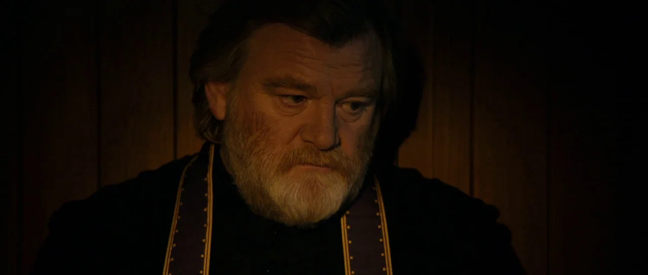 Calvary (2014) movie still: closeup — A dimly lit space with vertical wooden paneling serves as the backdrop. An older man in religious attire sits…