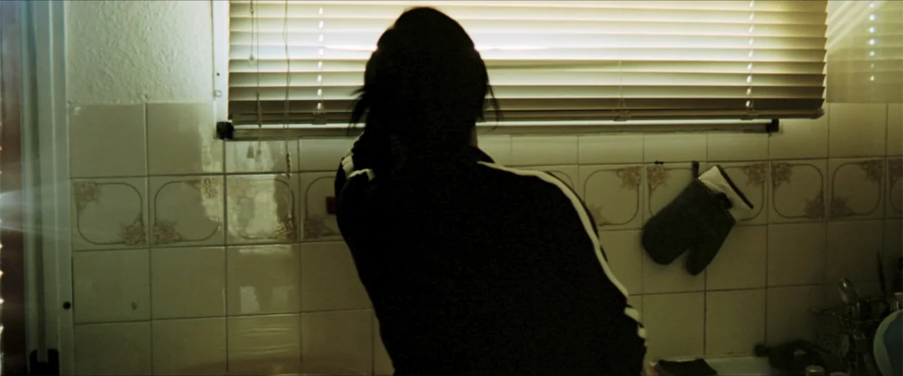 GOMORRAH (2023) movie still: medium — A residential room features light ceramic wall tiles and window blinds covering the aperture. A person with…