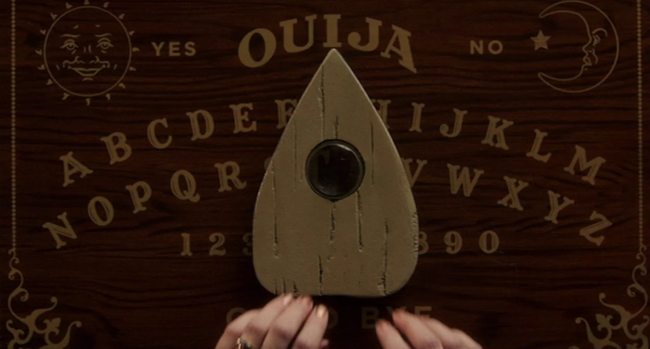 Ouija: Origin of Evil (2016) movie still: closeup — A dark, polished wooden surface features a printed alphabet board with sun and moon icons. A…