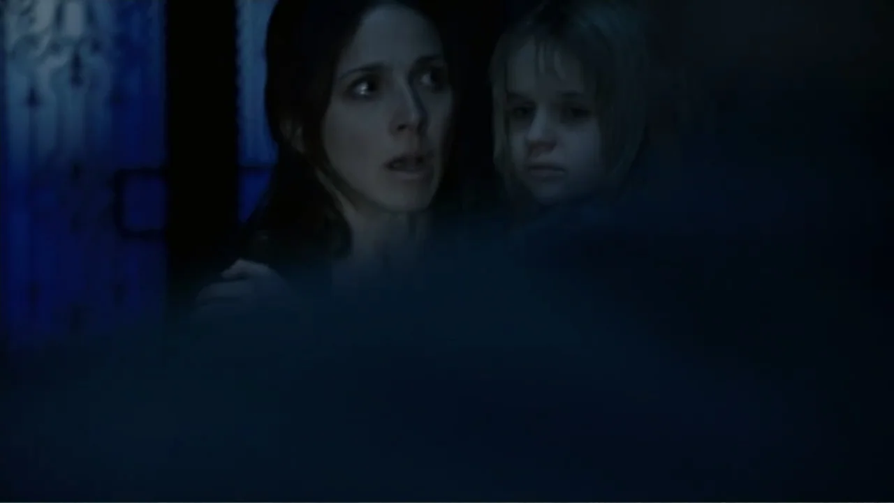 Quarantine (2008) movie still: medium — A dimly lit interior room at night shows two figures near a window. A woman embraces a child as both look…