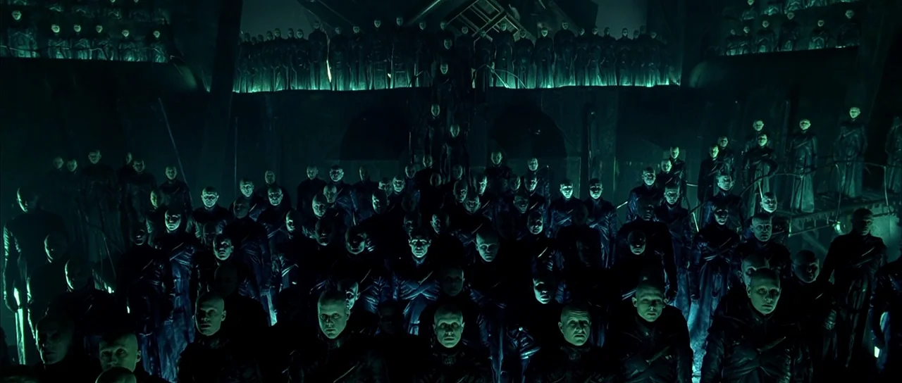 Dark City (1998) movie still: wide — A vast, dark amphitheater is filled with numerous pale, hairless humanoid figures in uniform. The crowd stands…