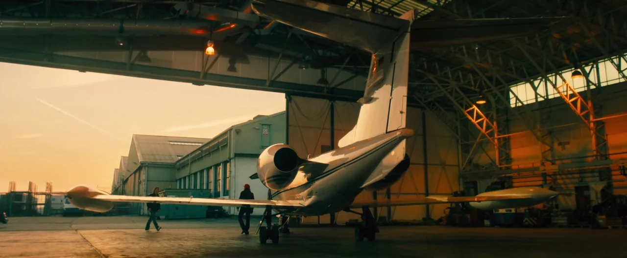 Rush (2013) movie still: wide — A cavernous aircraft hangar houses a private jet during the late afternoon. Two men walk toward the open hangar…
