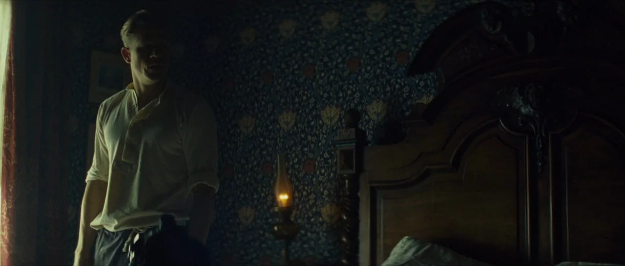 The Lost City of Z (2016) movie still: medium — A dimly lit bedroom features patterned floral wallpaper and a dark carved wooden headboard. A man in…
