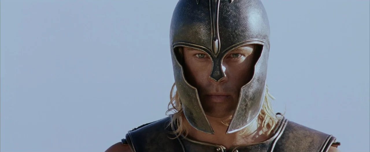 Troy (2004) movie still: closeup — An ancient warrior wearing a metallic helmet stands against a plain light-colored backdrop. The man faces forward…