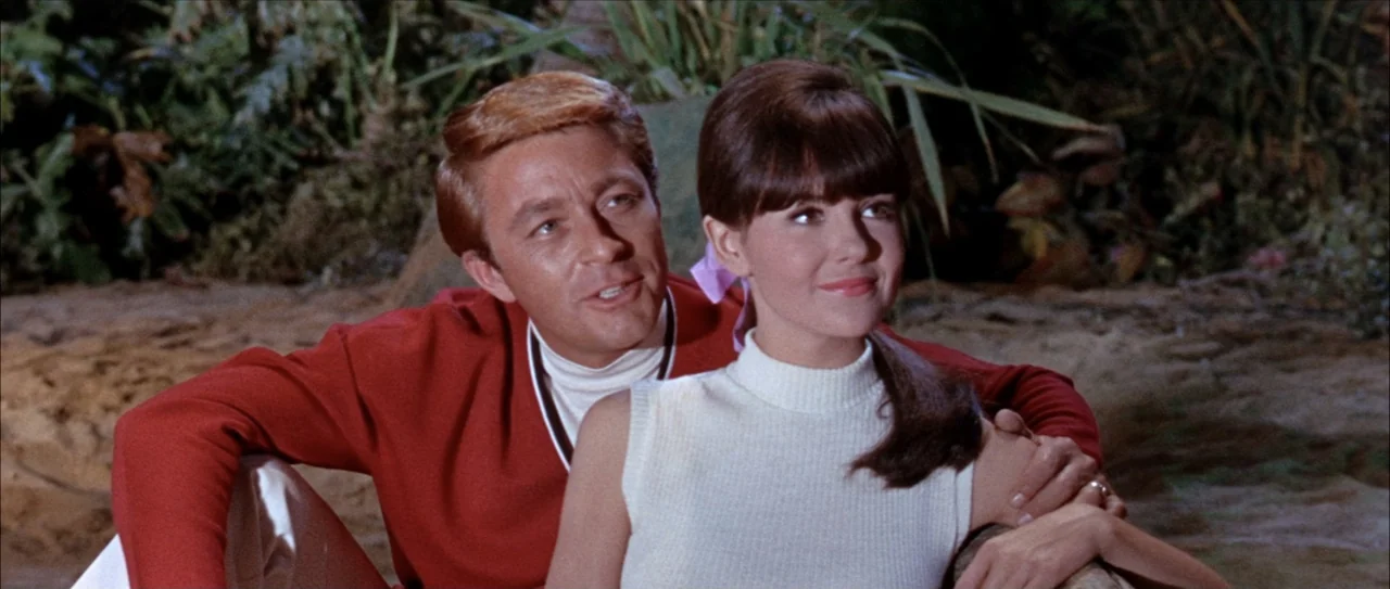 Clambake (1967) movie still: medium — A sandy beach environment with blurred tropical foliage in the background. A man and a woman sit together in…