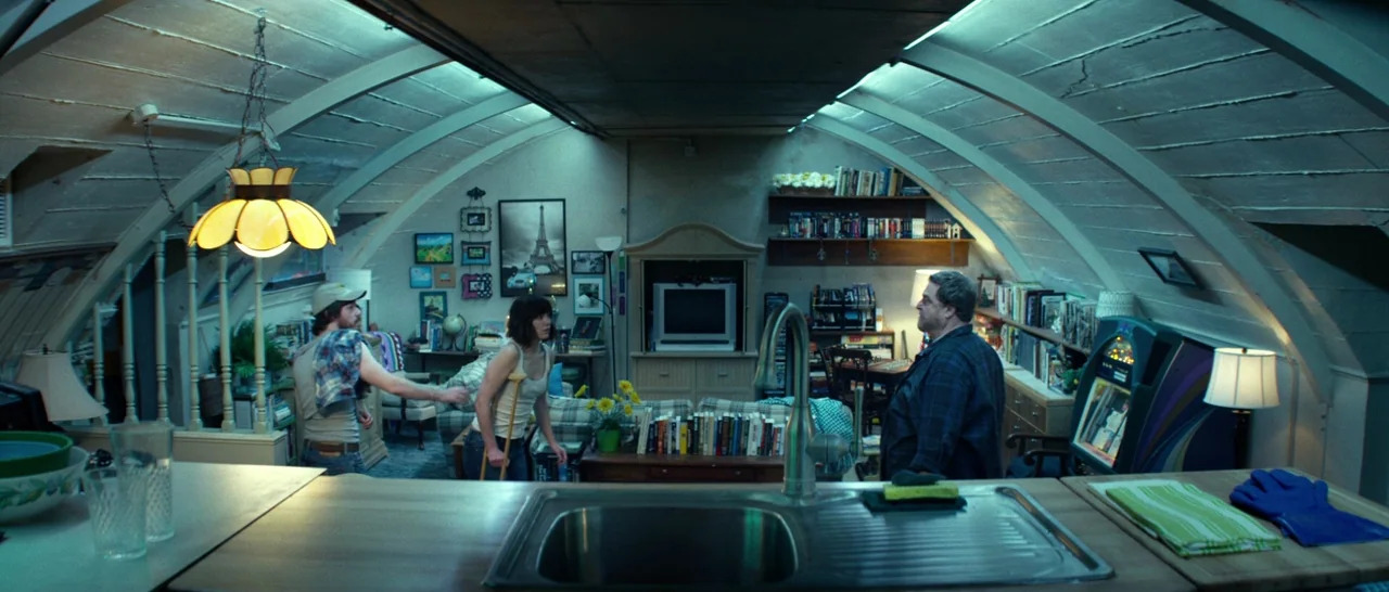 10 Cloverfield Lane (2016) movie still: wide — A subterranean bunker with curved concrete walls and residential furnishings including shelves of…