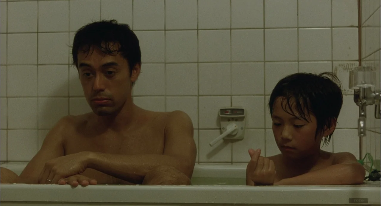 Still Walking (2008) movie still: medium — A square-tiled bathroom wall sits behind a white bathtub filled with water. A man and a young boy sit…
