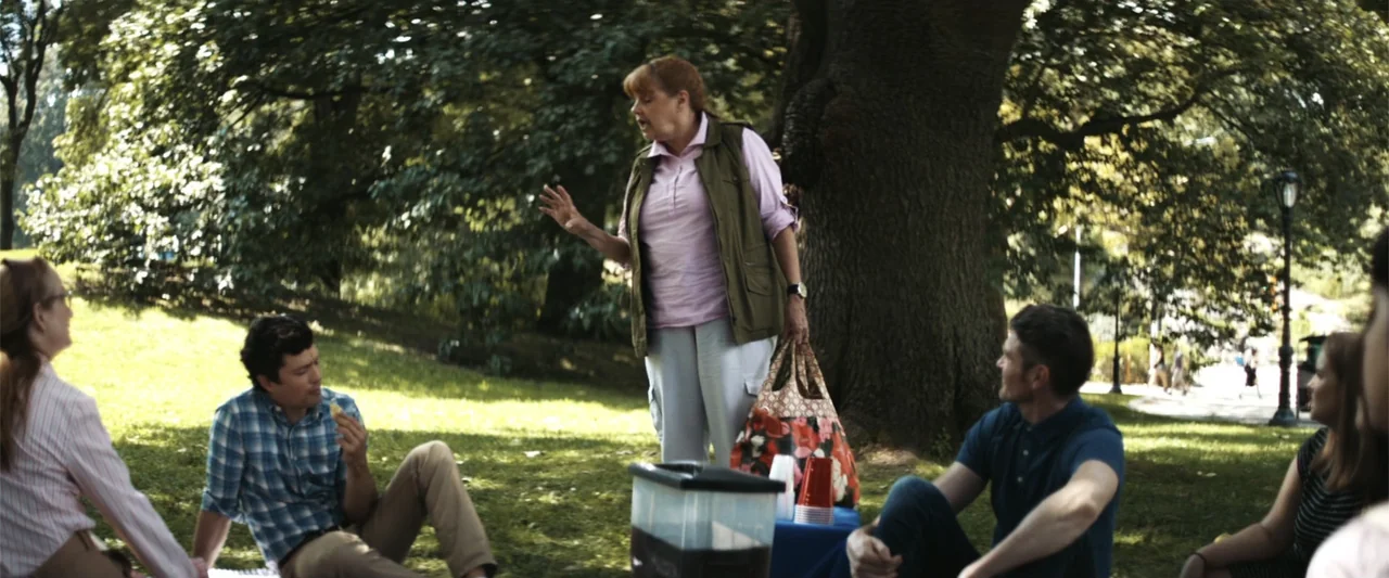 Mennonite's Bite (2023) movie still: fullbody — A group of people gathers on a grassy knoll under a large tree. A woman in a vest stands and…