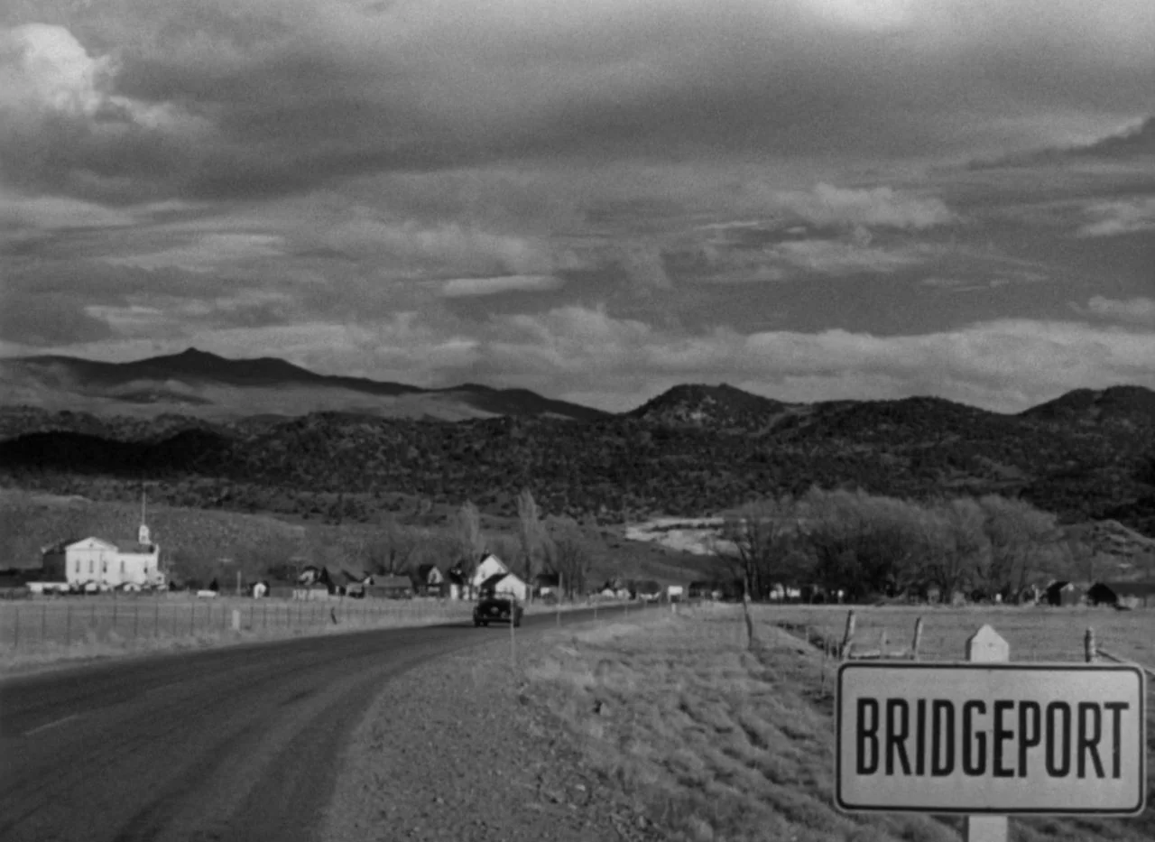 Out of the Past (1947) movie still: establishing — A rural landscape features a paved road leading toward a distant town with buildings. A sign…