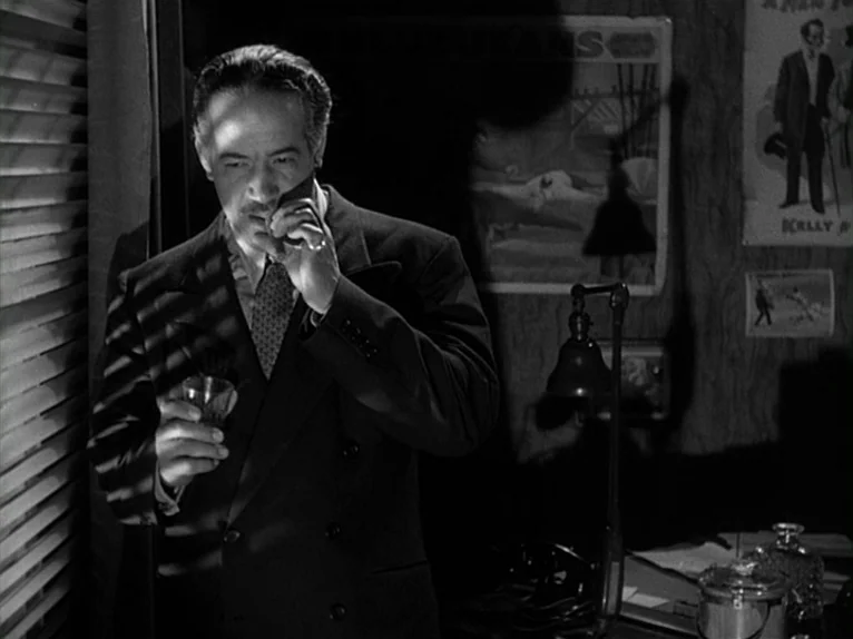 Killer’s Kiss (1955) movie still: medium — A dimly lit office features wood-paneled walls decorated with vintage posters. A middle-aged man stands…