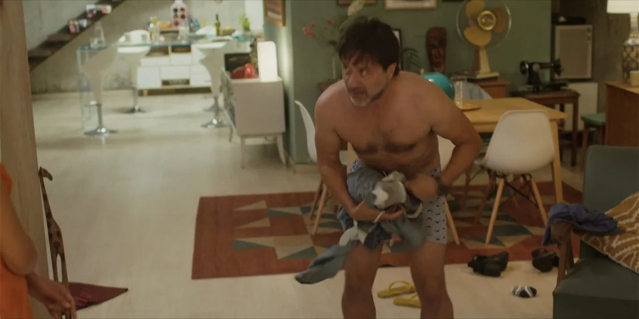 Instant Love (2020) movie still: medium — An open plan apartment features a dining table and wooden flooring with a rug. A middle-aged man stands in…