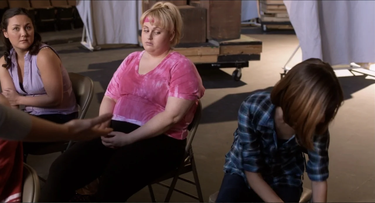 Pitch Perfect (2012) movie still: medium — A utilitarian rehearsal space features folding chairs and rolling equipment carts in the background.…