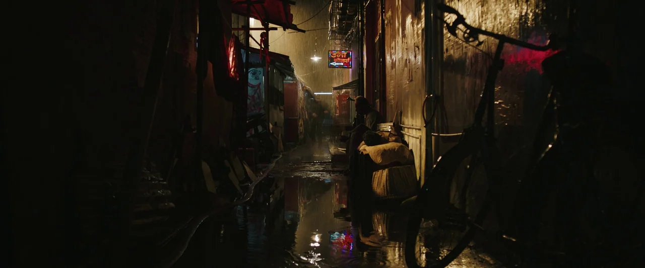 Monkey Man (2024) movie still: wide — A narrow rain-slicked urban path is flanked by weathered walls covered in posters. A dark bicycle silhouette…