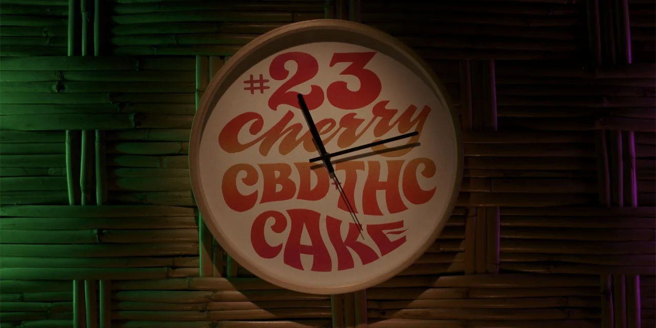 Sitting in Bars with Cake (2023) movie still: closeup — A circular wall clock with retro-style typography is mounted on a brown bamboo wall. Green…