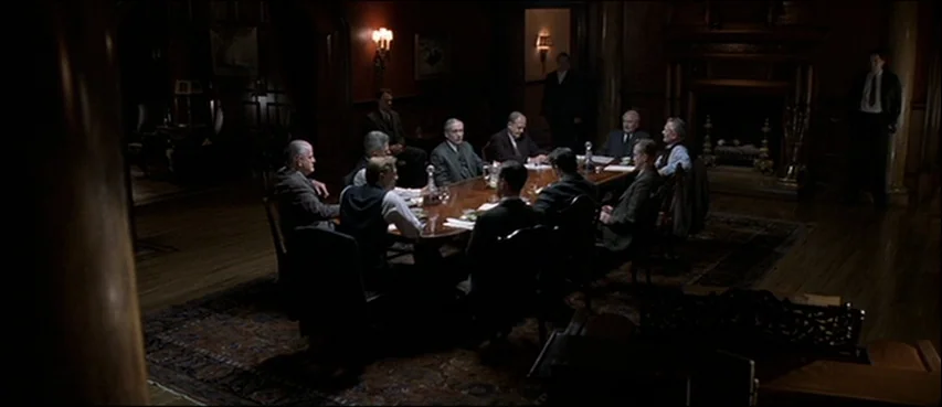 Road To Perdition (2002) movie still: wide — A dark, wood-paneled room with a large conference table surrounded by ten seated men. The group holds a…