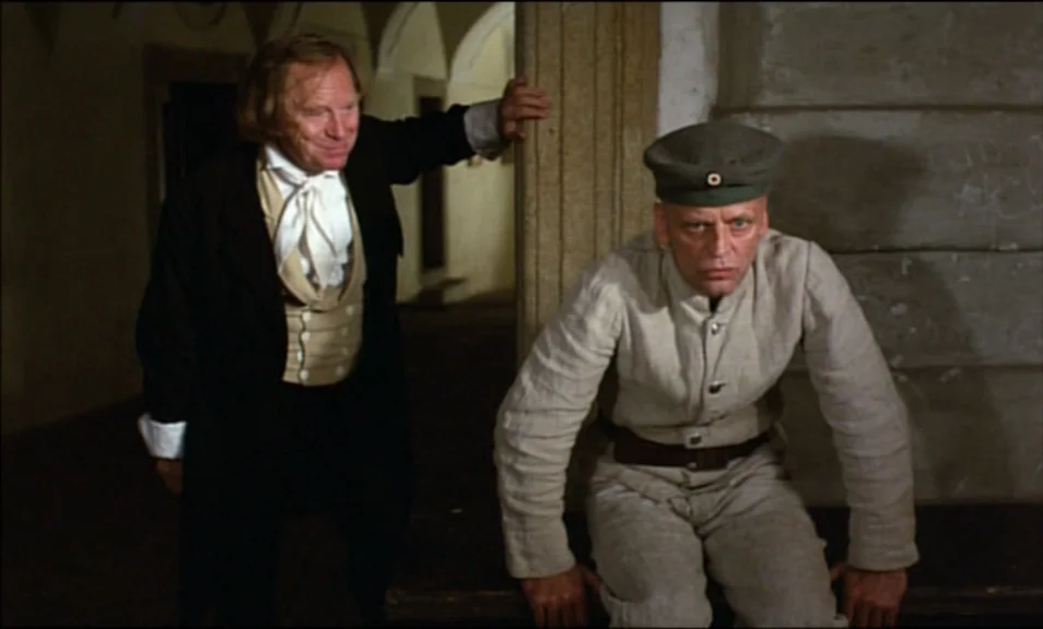 Woyzeck (1979) movie still: medium — A dimly lit indoor corridor with stone walls and arched doorways in the background. A man in formal…