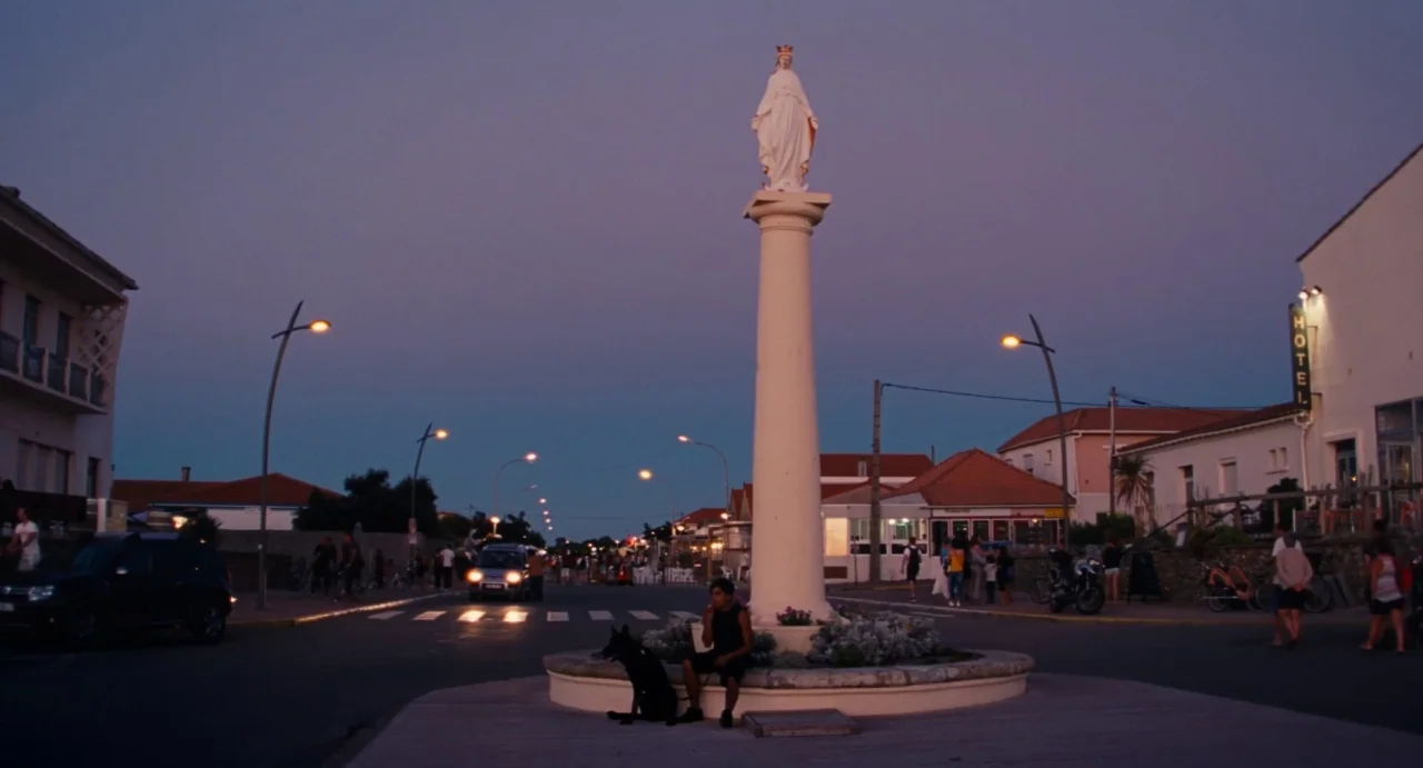Ava (2017) movie still: wide — A town square at dusk features a tall pillar topped with a white statue. A young person sits at the base of the…