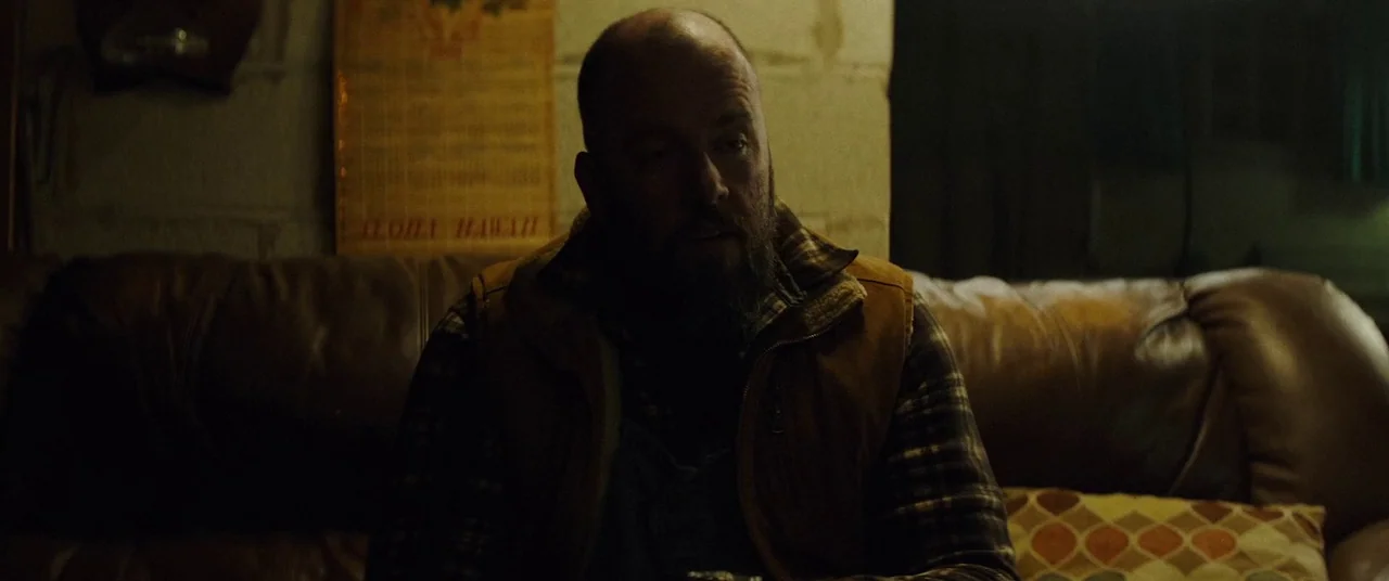 Broadcast Signal Intrusion (2021) movie still: medium — A man sits on a brown leather couch in a dimly lit, cluttered room. A vintage wall calendar…
