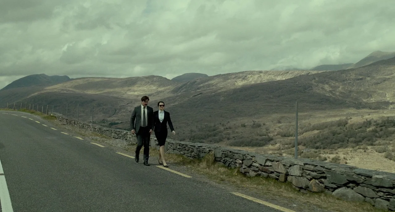 The Lobster (2015) movie still: wide — A man and a woman in formal business attire walk along a remote road. They pass a stone wall with mountains…
