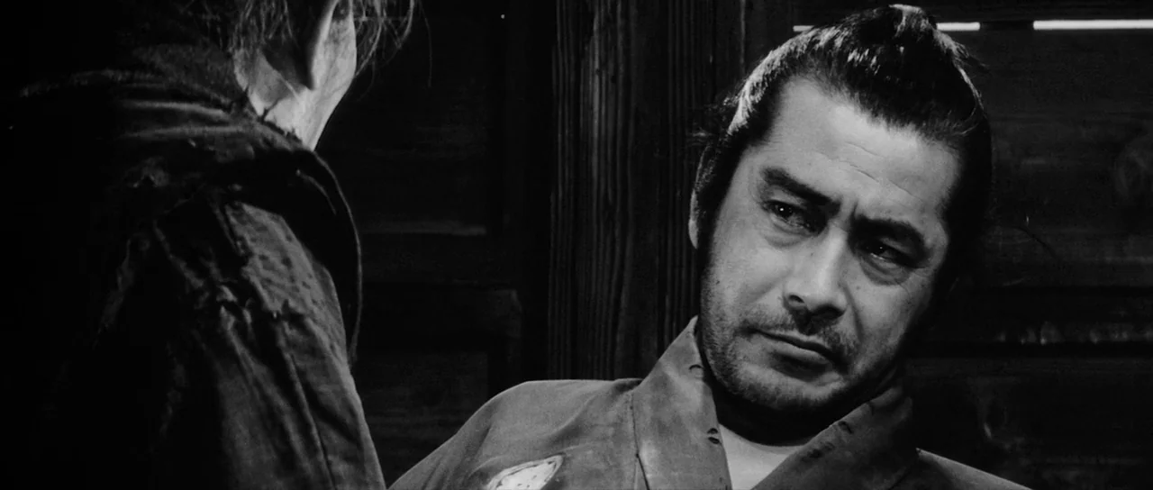 Yojimbo (1961) movie still: medium — A dark, dimly lit wooden interior with rough plank walls. A man with a topknot listens while another person…