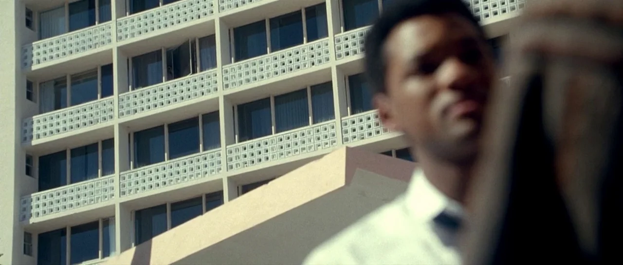 Ali (2001) movie still: medium — A modern multi-story apartment building features repeating white geometric balconies. An out-of-focus man stands in…