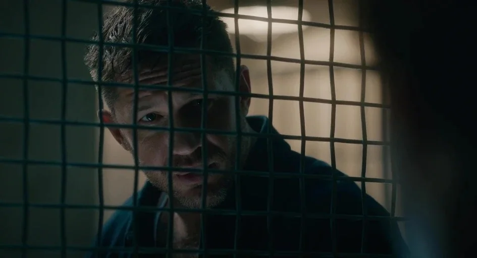 Venom Let There Be Carnage (2021) movie still: closeup — A man stands behind a metal security grid in a dimly lit room. He peers through the square…