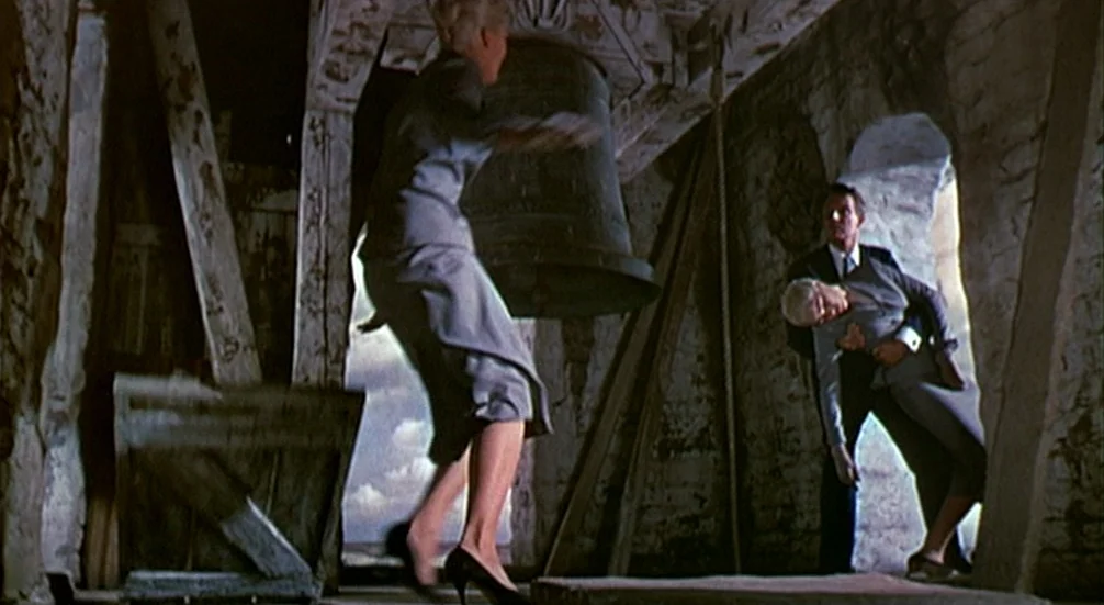 Vertigo (1958) movie still: wide — A stone bell tower interior features wooden support beams and a large iron bell. A man holding an unconscious…