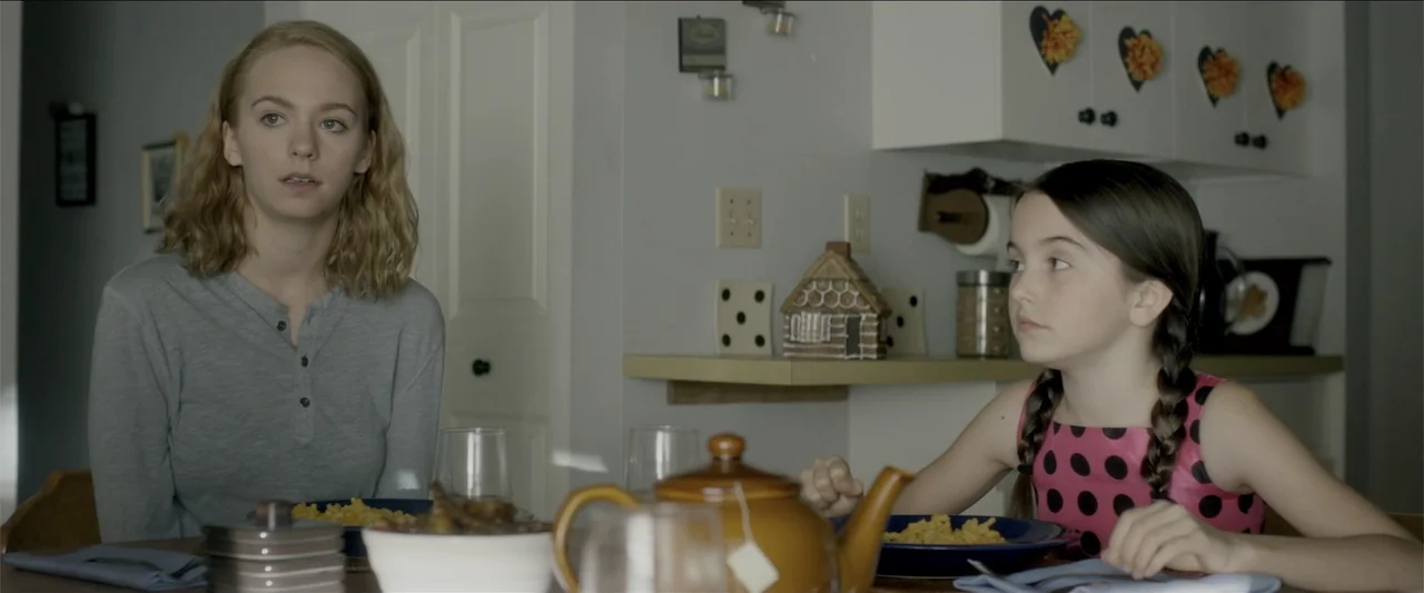 The Hollow Child (2018) movie still: medium — A residential room features light gray walls and a dining table with place settings. Two females sit…