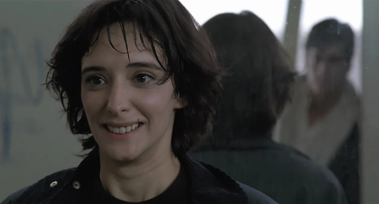 Thesis (1996) movie still: closeup — A woman stands before a mirror in a brightly lit, sterile space. She gazes at her own reflection while wearing…