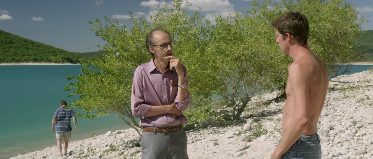 Stranger By The Lake (2013) movie still: medium — A rocky lakeshore with turquoise water and green hills in the distance. An older man in a dress…