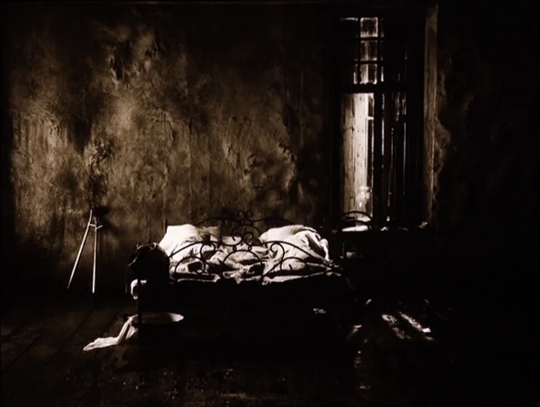 Stalker (1979) movie still: wide — A dilapidated, dimly lit space with dark textured walls and a wrought-iron bed frame. A single window allows…